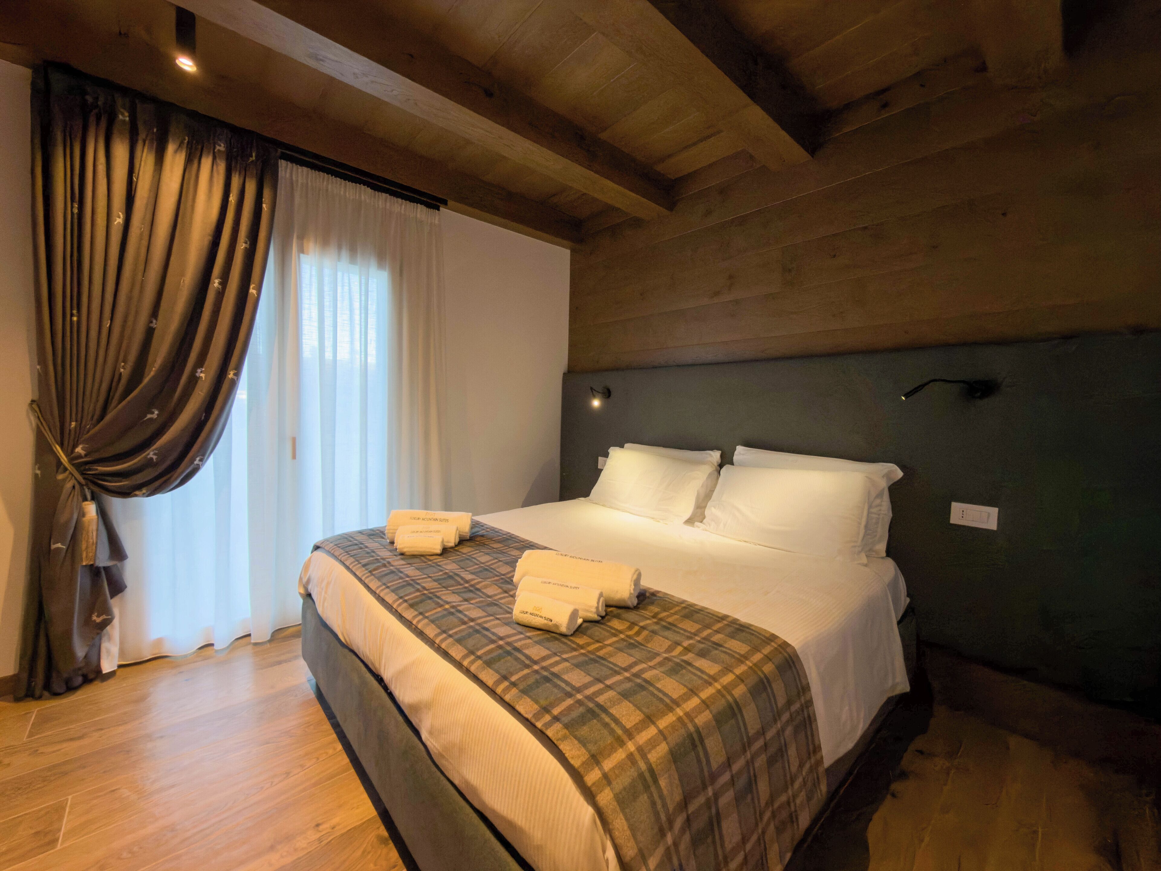 A hotel room with a wooden ceiling, a bed with white linens, a plaid blanket, and a window with curtains.