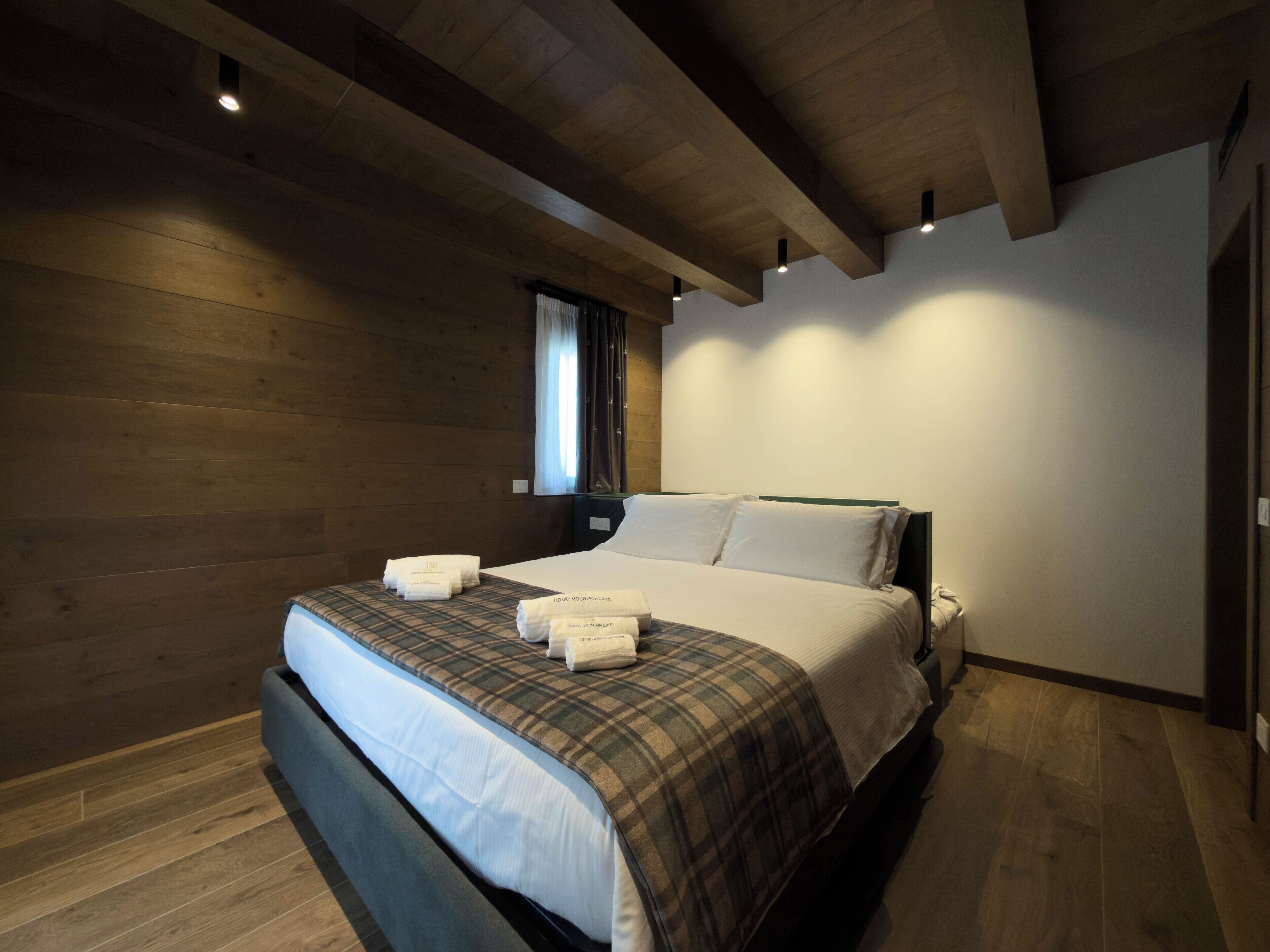 A modern hotel room with a wooden floor, a large bed with a plaid bedspread, and a window with curtains.