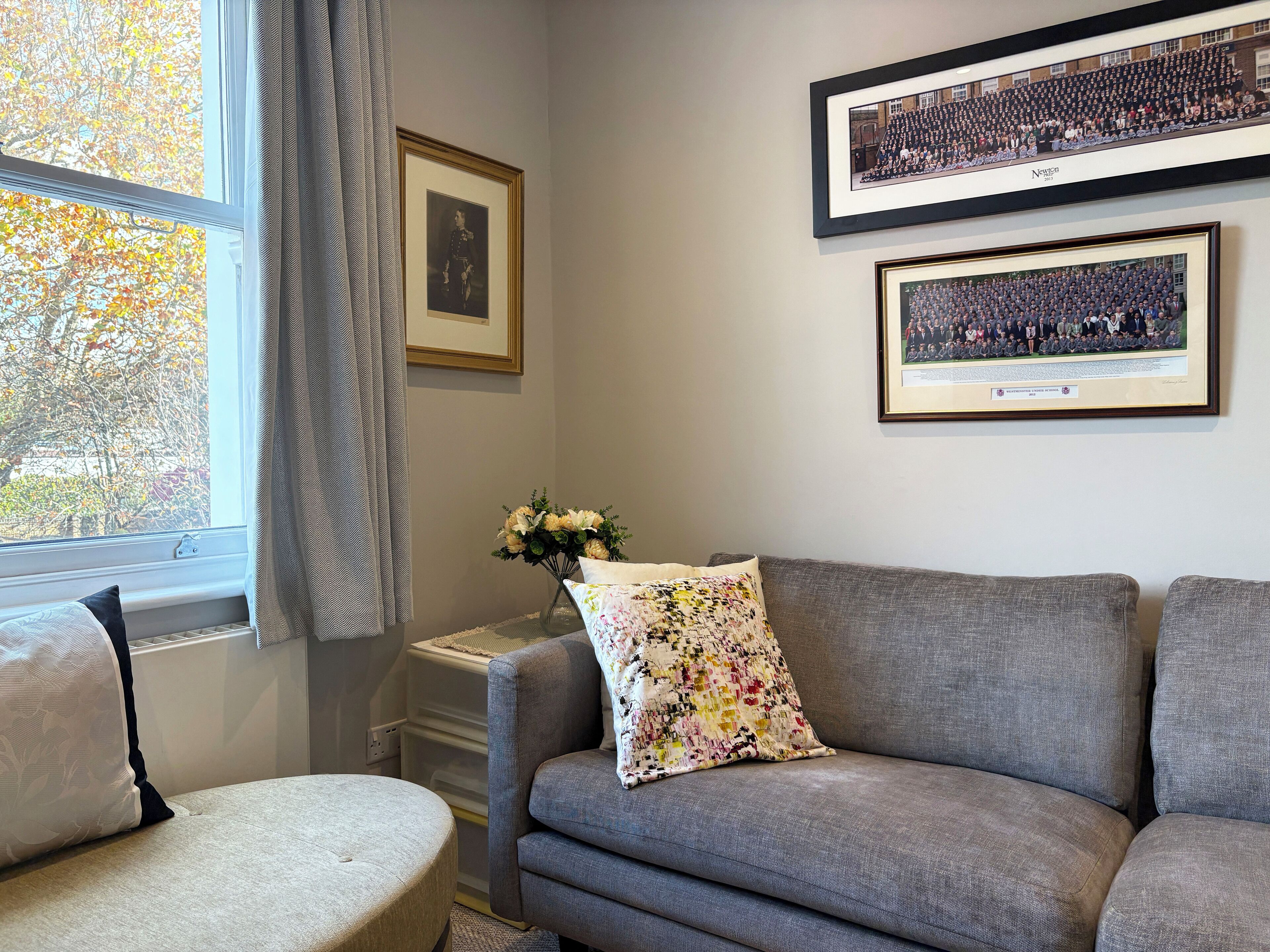 A living room with a grey sofa, a small round ottoman, a floral pillow, and a framed photograph on the wall.