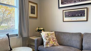 A living room with a grey sofa, a small round ottoman, a floral pillow, and a framed photograph on the wall.