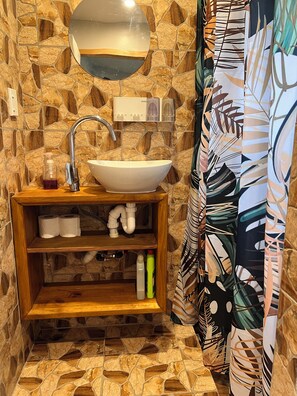 Un baño con pared de piedra, un espejo redondo, un lavabo blanco, una estantería de madera con artículos de aseo, y una cortina de ducha estampada.