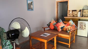 Een kamer met een houten tafel en stoelen, een ventilator, een kleine koelkast en een deur naar een andere kamer.