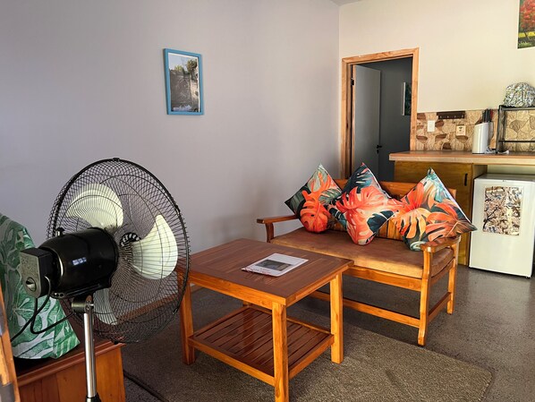 Una habitación con una mesa y sillas de madera, un ventilador, un refrigerador pequeño y una puerta que da a otra habitación.