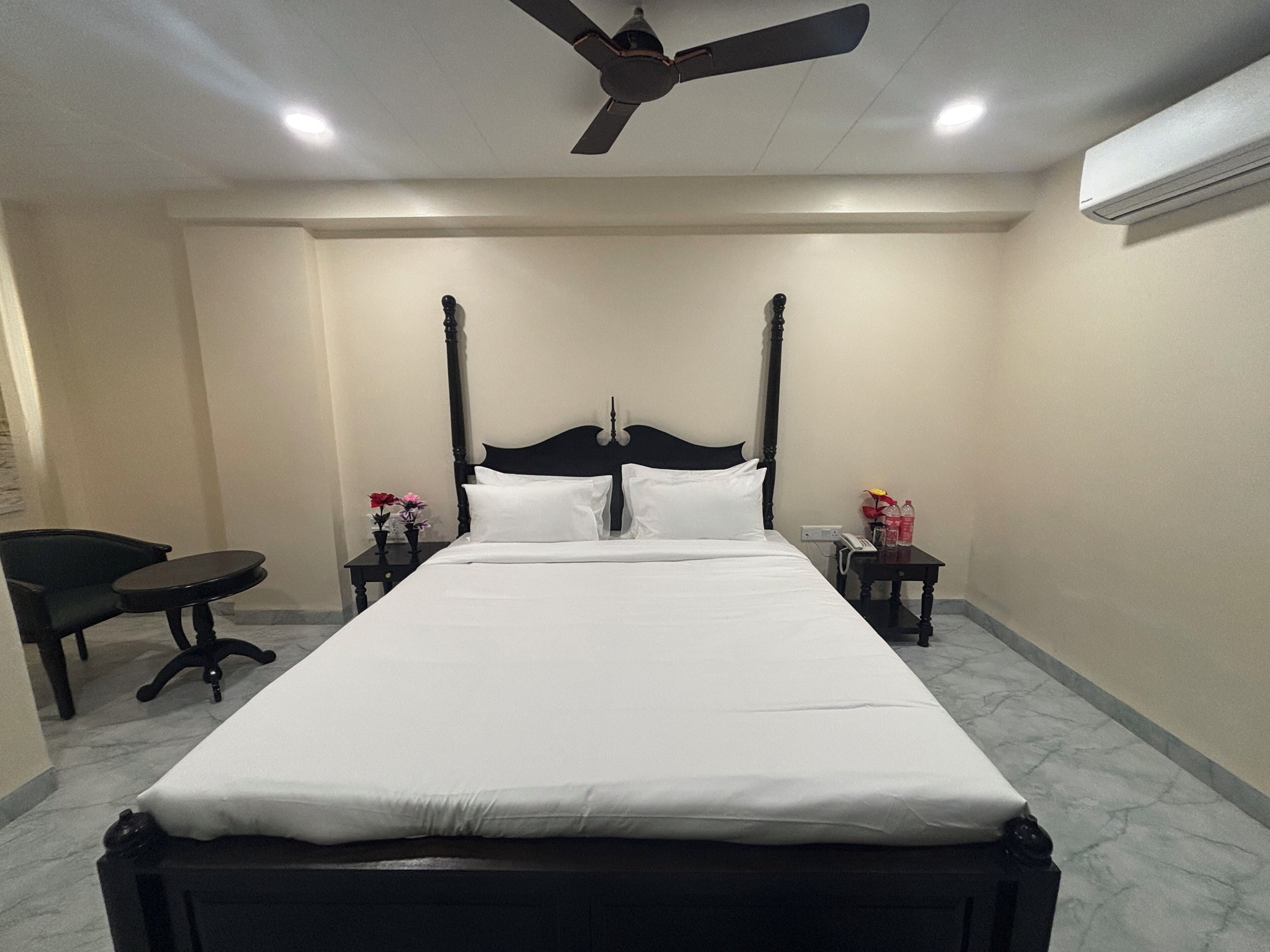A neatly made bed in a hotel room with a ceiling fan, a small table with a chair, and a nightstand with a phone and flowers.