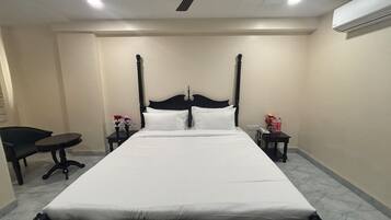 A neatly made bed in a hotel room with a ceiling fan, a small table with a chair, and a nightstand with a phone and flowers.