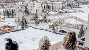 Trois personnes vêtues de blouses blanches se tiennent sur un balcon qui donne sur un paysage enneigé, avec des bâtiments et une piste de ski.
