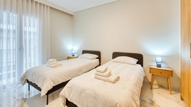 A hotel room with two single beds, a nightstand with a lamp, and a large window with sheer curtains. - Thessaloniki Riviera Modern Stay by ΜΡS (Kalamaria)