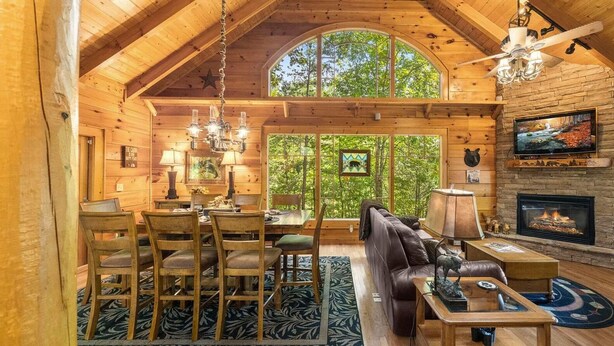 A cozy cabin interior with a high ceiling, wooden walls, a fireplace, a television, and a dining area with chairs and a table. - Sleeps 10, 3 BR + Loft - Hot Tub, Fire Pit, Beautiful Cozy Cabin (Sevierville)