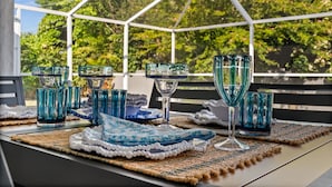 Mesa dispuesta con cristalería azul y platos blancos, sobre un mantel tejido, bajo una cubierta de vidrio con vista a árboles.