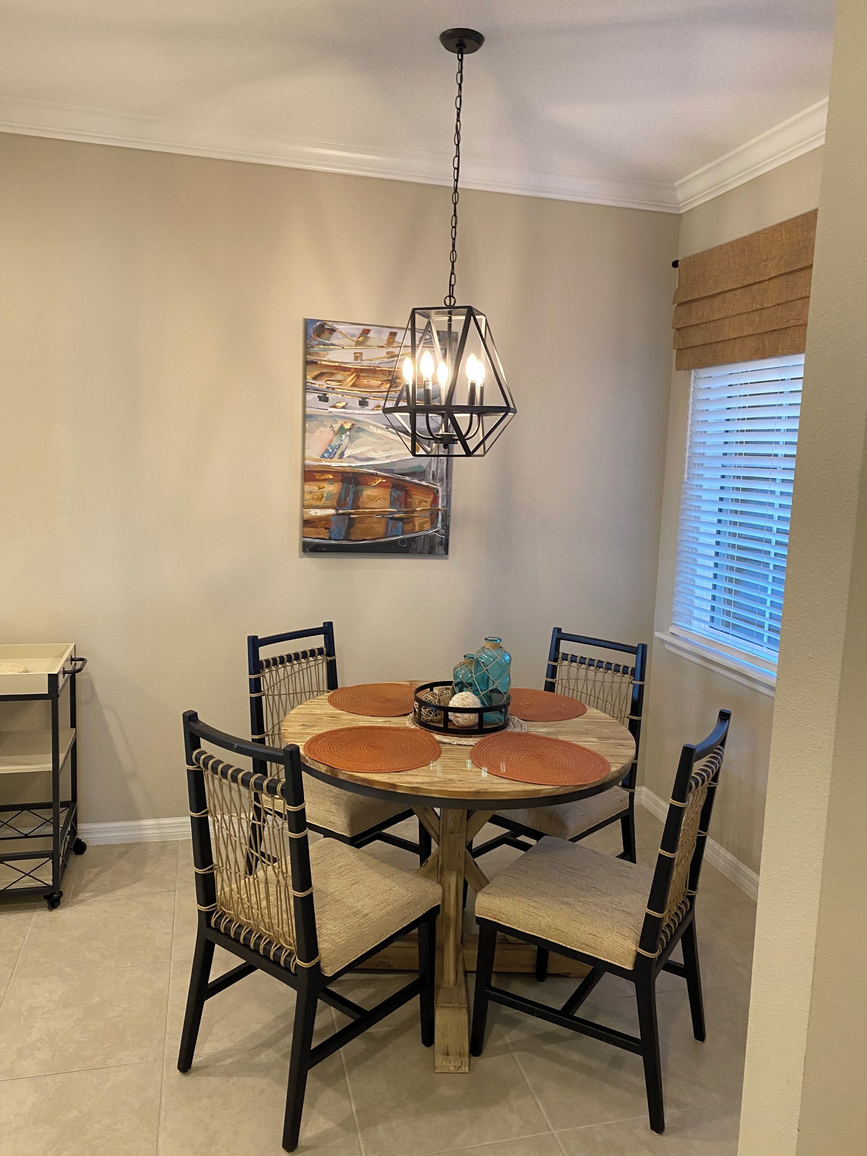 A dining area with a round wooden table, four chairs, a hanging light fixture, and a window with blinds.