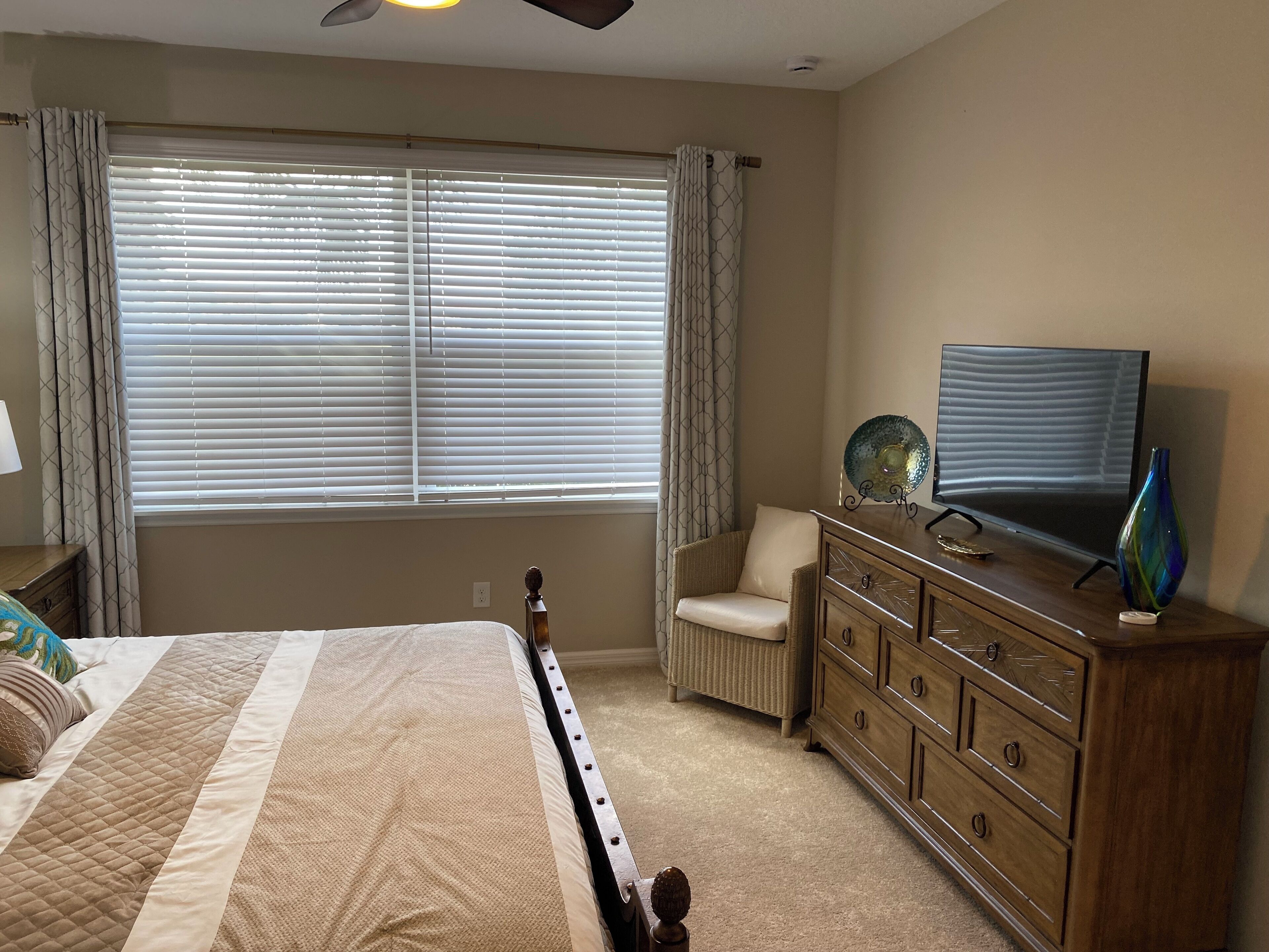 Une chambre à coucher comprenant un lit, une commode, une chaise, un téléviseur et une fenêtre avec des stores.