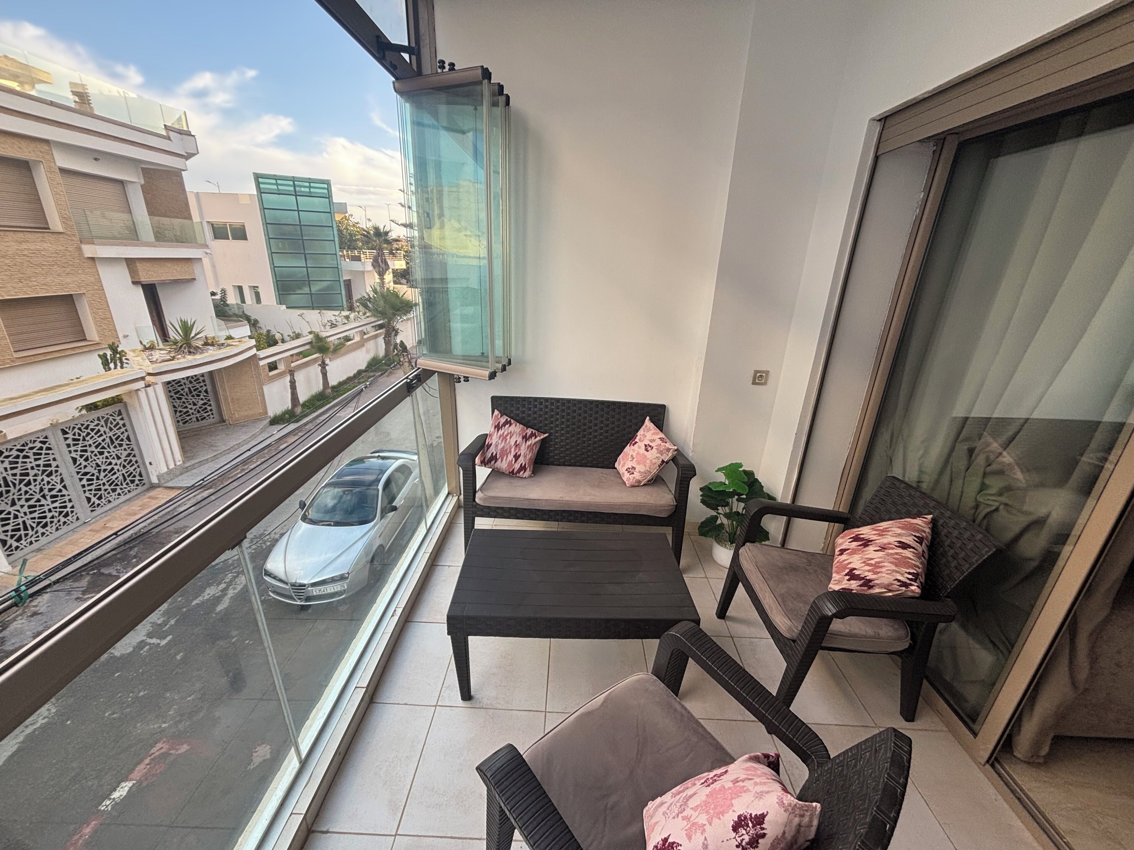 A balcony with a sofa and chairs, a view of a street with a car, and a building in the background.