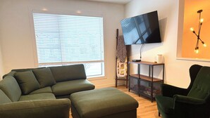 A living room with a sectional sofa, an ottoman, a single armchair, a TV on the wall, and a small table with a lamp. - Townhouse - 2 bedrooms, both ensuites! (Red Deer)