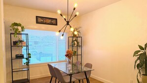 A dining area with a glass table, four chairs, a chandelier, and two black shelving units with plants. - Townhouse - 2 bedrooms, both ensuites! (Red Deer)