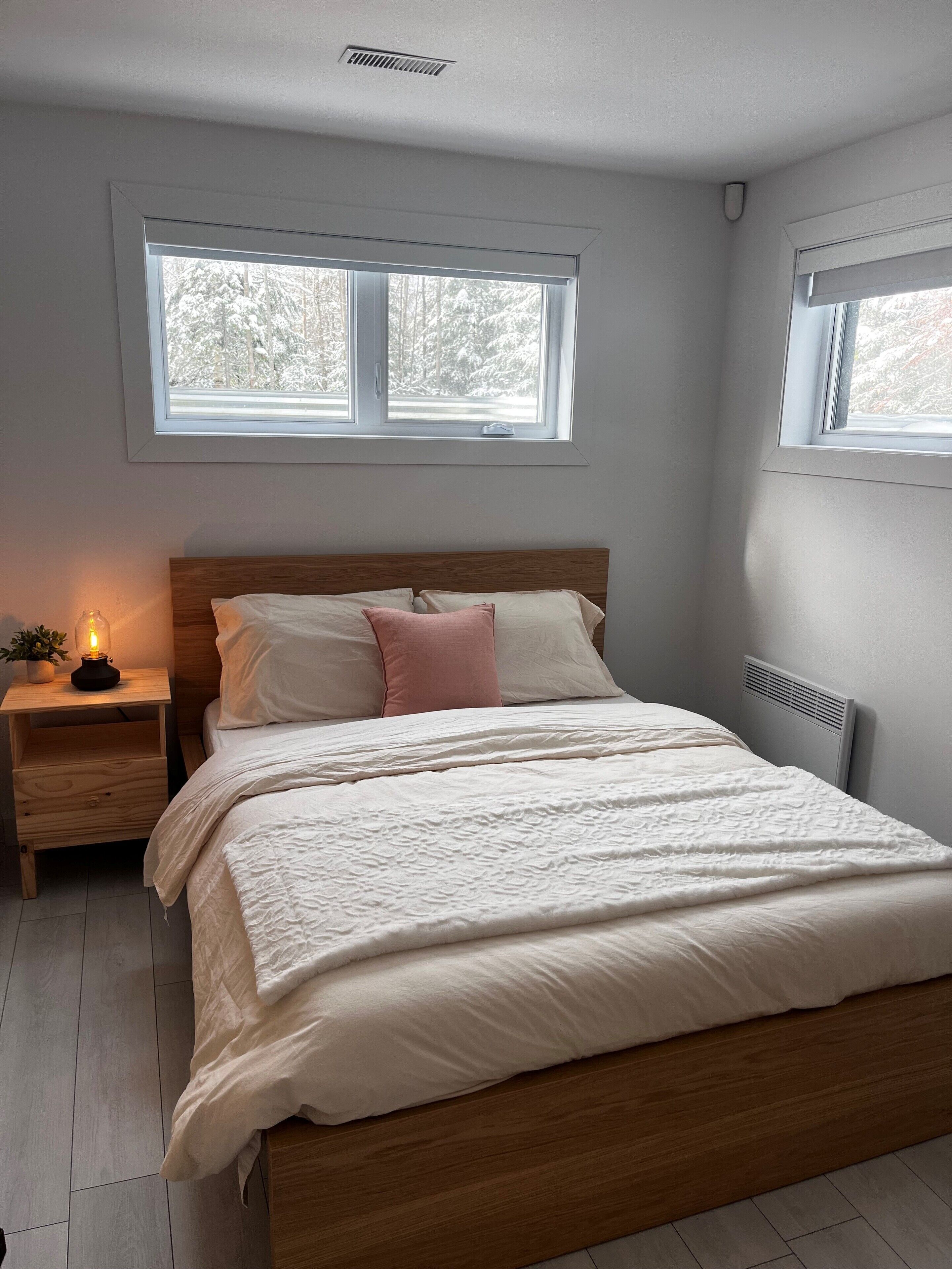 Un dormitorio con cama de madera, ropa de cama blanca, una mesita de noche con lámpara y una ventana con vistas a árboles cubiertos de nieve.