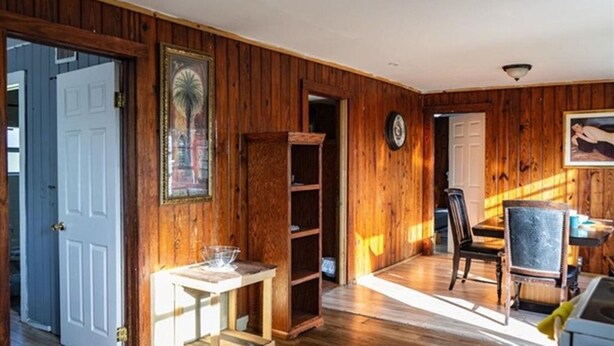 Una habitación con paredes de madera, una puerta blanca, una estantería, una mesa de comedor con sillas y un cuadro en la pared.