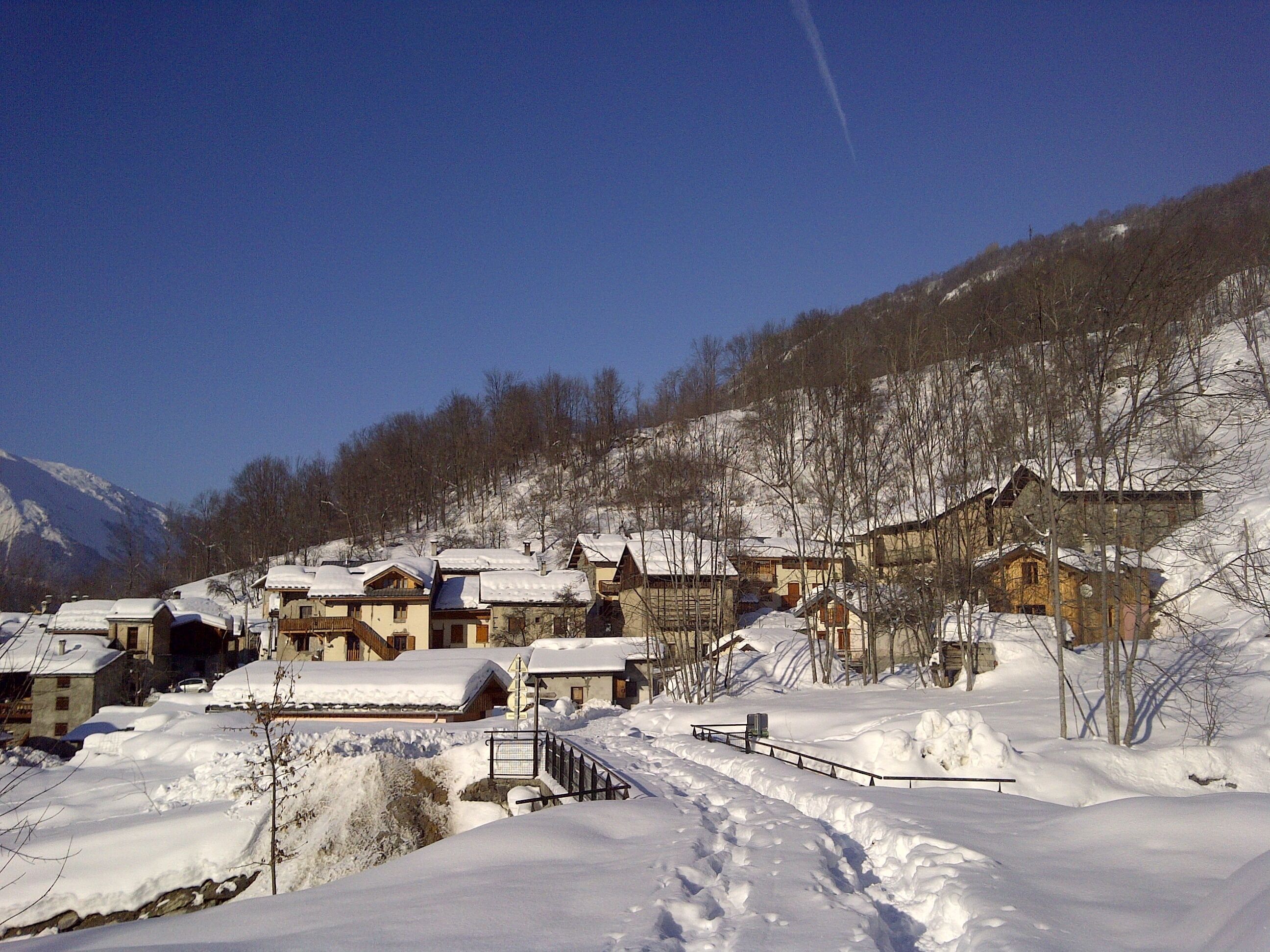 A snow-covered village with traditional wooden houses, a small bridge, and a mountain backdrop.
