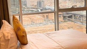 Una cama con ropa de cama blanca, cabecera marrón y vista de un paisaje urbano a través de ventanas amplias.