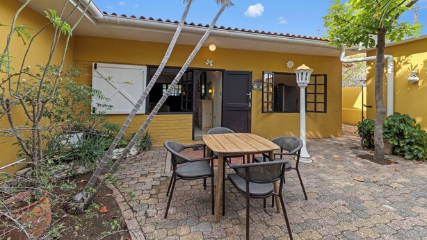 Um edifício amarelo com telhado de azulejos, uma mesa de madeira com quatro cadeiras e uma pequena área de jardim com plantas e uma árvore.