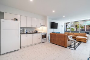 A modern kitchen with white cabinets, a built-in oven, and a refrigerator. There is a brown leather sofa and a small round table in the living area. - BeachSide 9th Residences (Fort Lauderdale)