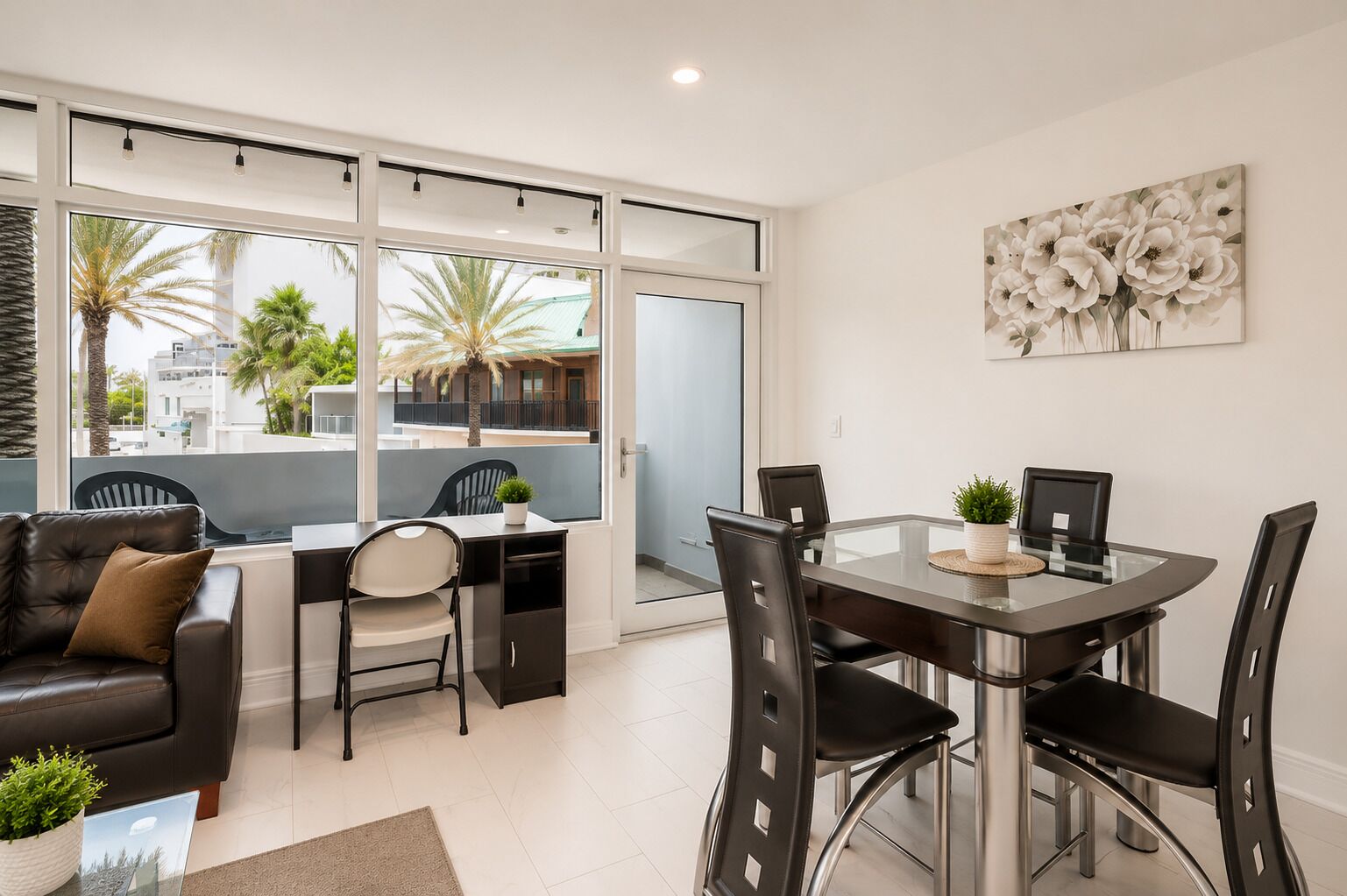 A modern living room with a glass dining table, black chairs, a leather sofa, and a large window with a view of palm trees and buildings.