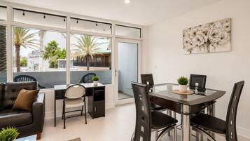 A modern living room with a glass dining table, black chairs, a leather sofa, and a large window with a view of palm trees and buildings.