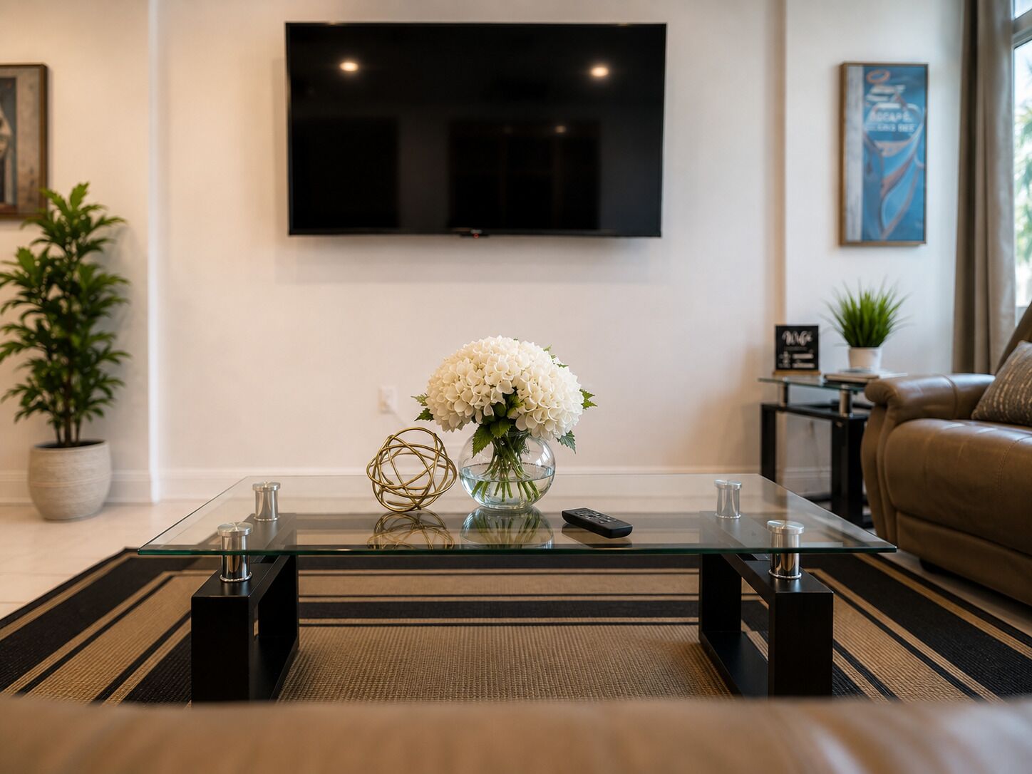 A modern living room with a flat-screen TV mounted on the wall, a glass coffee table with a vase of flowers, and a leather sofa.
