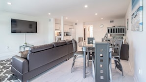 A modern living room with a grey sofa, a dining table with chairs, a flat-screen TV mounted on the wall, and a kitchen area in the background. - BeachSide 9th Residences (Fort Lauderdale)