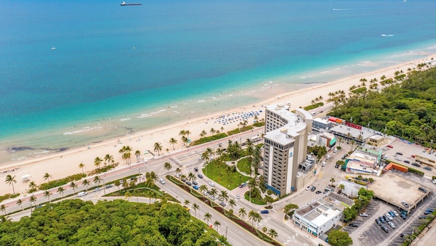 A coastal cityscape with a beach, palm trees, and a large building with a parking lot. - BeachSide 9th Residences (Fort Lauderdale)