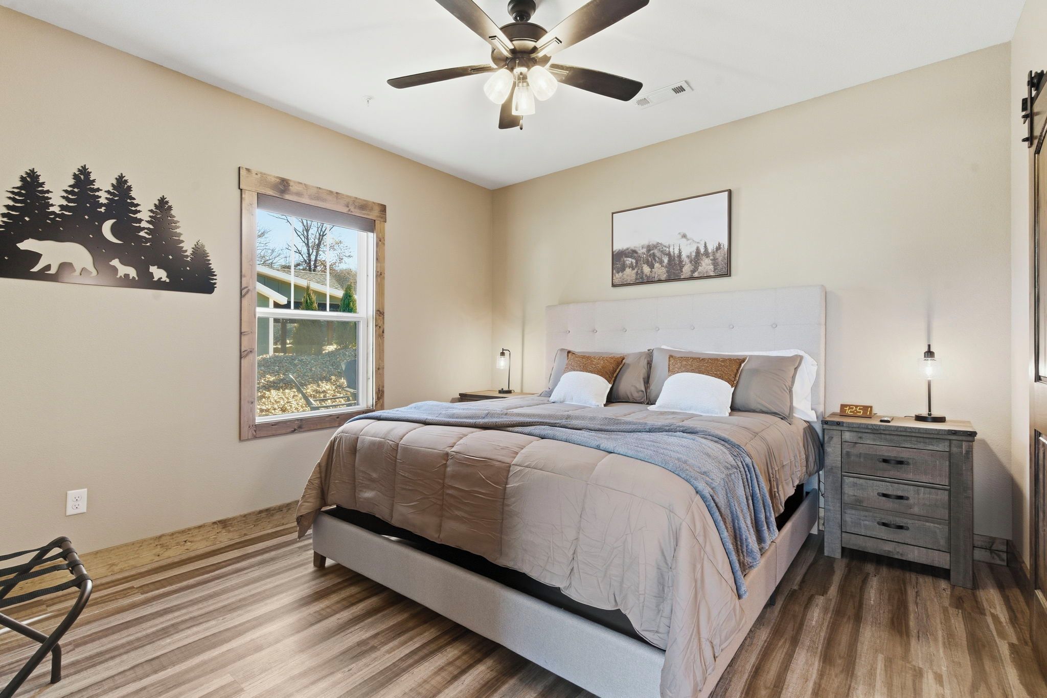 A bedroom with a bed, a nightstand, a dresser, a ceiling fan, a window with a view, and wall art.