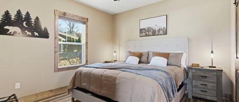 A bedroom with a bed, a nightstand, a dresser, a ceiling fan, a window with a view, and wall art.