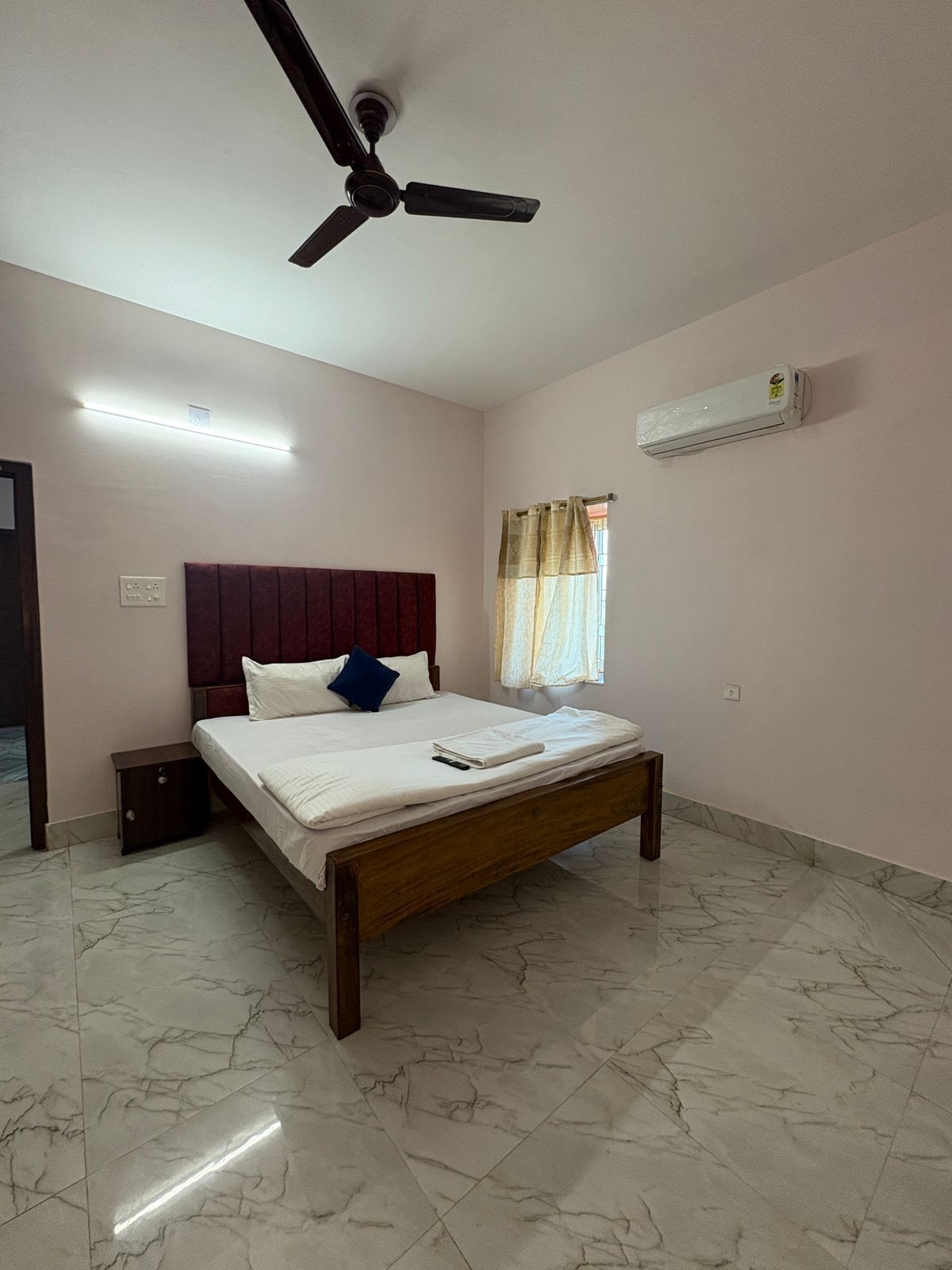 A bedroom with a bed, a ceiling fan, an air conditioner, a nightstand, and a window with curtains.