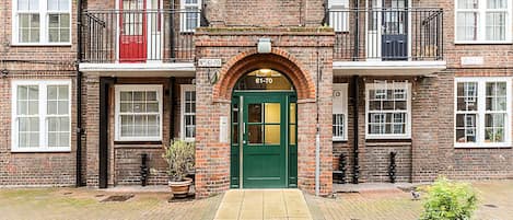 A brick building with a green entrance door, multiple windows, and a small balcony.