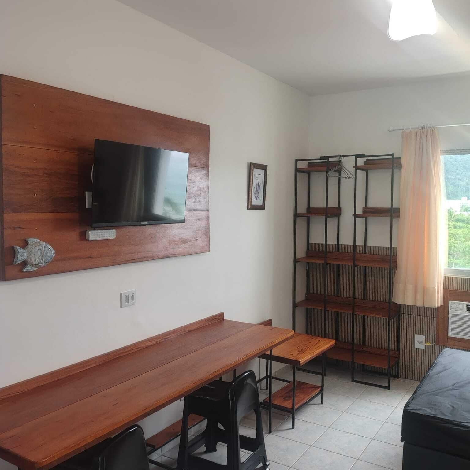 A room with a wooden desk, a TV mounted on the wall, a bookshelf, and a window with curtains.