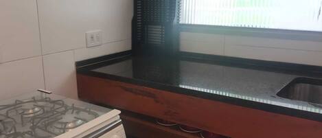 Una cocina con estufa a gas, encimera negra, ventana con persianas y pared de azulejos blancos.