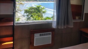 Una habitación con un ventanal grande que da a una playa y montañas. Hay un estante de madera y un equipo de aire acondicionado instalado en la pared.