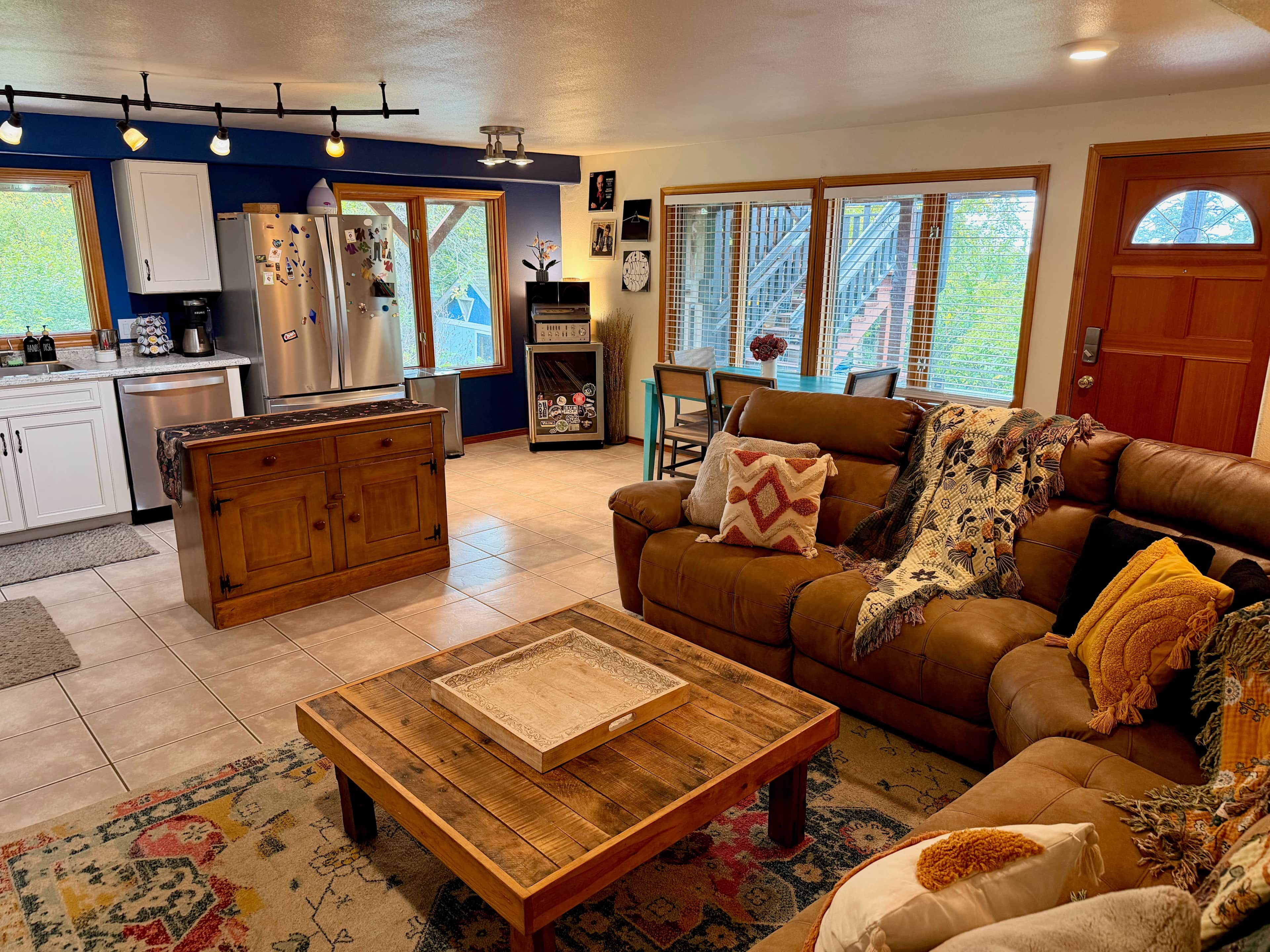 A living room with a brown leather sofa, a wooden coffee table, and a kitchen area with a refrigerator and cabinets.