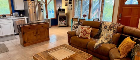 A living room with a brown leather sofa, a wooden coffee table, and a kitchen area with a refrigerator and cabinets.