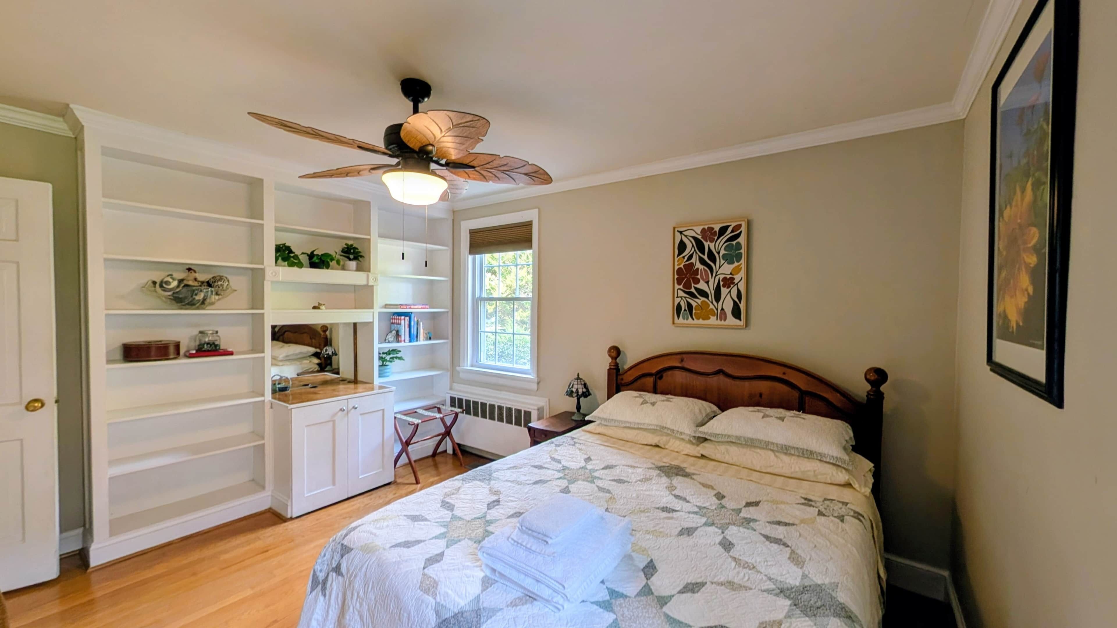 Un dormitorio con cama, ventilador de techo, estanterías empotradas, una ventana y obras de arte en la pared.