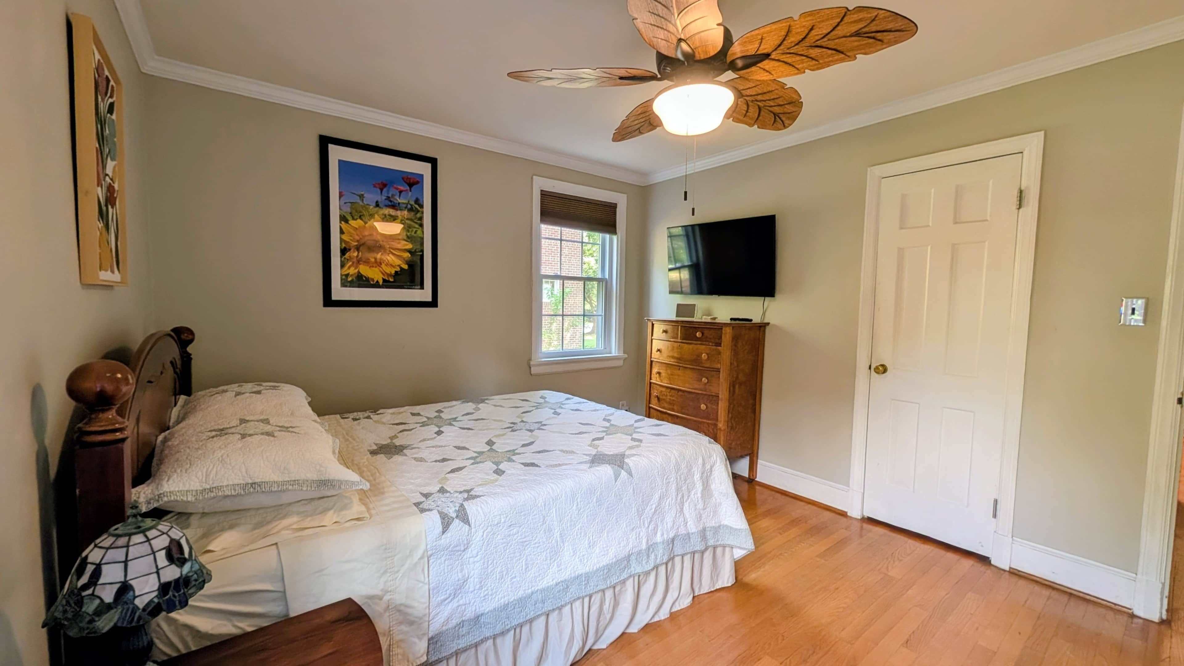 A bedroom with a bed, a ceiling fan, a dresser, a television, and a door.