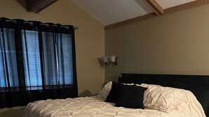 Un dormitorio con cama, ventana con cortinas, lámpara de pared y techo con vigas de madera.