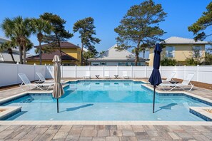 A rectangular swimming pool with a tiled edge, surrounded by lounge chairs and umbrellas, with a white fence and trees in the background.
