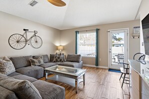 A modern living room with a sofa set, a glass coffee table, a bicycle mounted on the wall, and a view through a door to the outside.