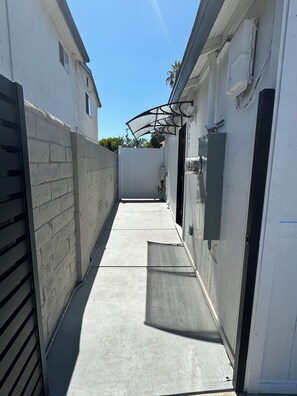 Ein schmaler Betonweg zwischen Gebäuden mit weißer Fassade und einem Metallgitter links.