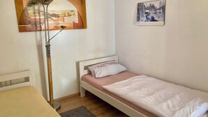 A small, single-bed room with a wooden floor, a bedside table, a floor lamp, and two wall-mounted pictures.