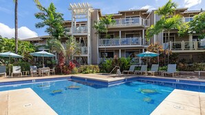 Piscina con sillones de descanso y sombrillas, rodeada de palmeras y un edificio de varios pisos con balcones.