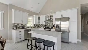 Uma cozinha moderna com armários brancos, uma ilha central com banquetas, eletrodomésticos de aço inoxidável e uma parede de proteção revestida de azulejos.