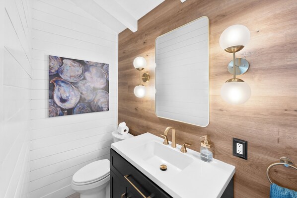 A modern bathroom with a wooden wall, a white sink with gold fixtures, a large mirror, and a painting of abstract shapes.