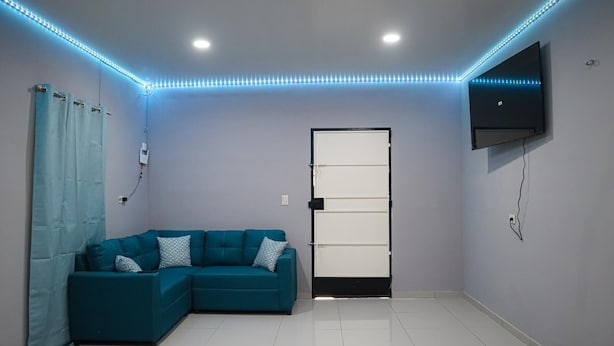 Une pièce comprenant un canapé d’angle de couleur turquoise, une porte blanche, un téléviseur à écran plat fixé au mur et un éclairage décoratif à LED le long du plafond.