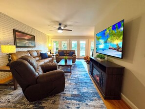 Una sala de estar con sofás de cuero, un televisor de pantalla plana, un ventilador de techo y un ventanal grande.